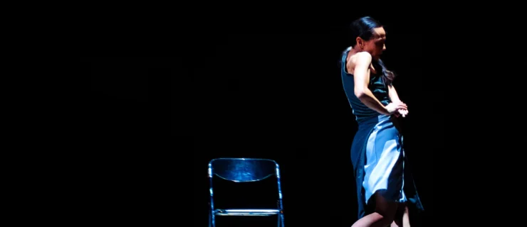 Robyn Mineko Williams dances alone in a spotlight beside an empty folding chair. She is captured in profile, shoulder rising to meet her cheek as she twists and pushes her hands down from hip height. Her blue dress twists above pretzeled lower legs. She is barefoot.