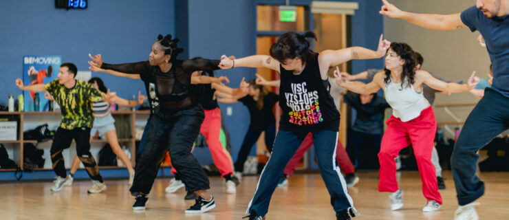 Nina Flagg (in all black) leading a hip hop class to a large group of students in a studio. They extend their arms side while tapping their right foot out