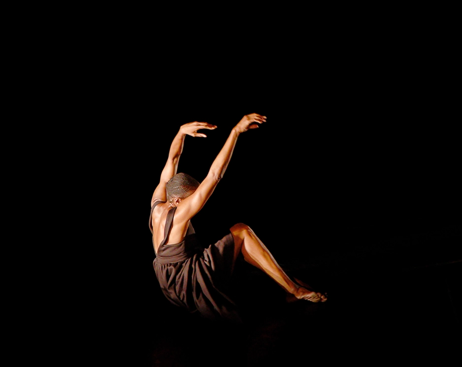 Okwui Okpokwasili seems almost to float as she sits on the floor in a dark space. She is twisted away from the camera, face tucked against her shoulder as her arms drift overhead. Her knees pull up toward her chest, feet dragging along the floor. She wears a brown dress.