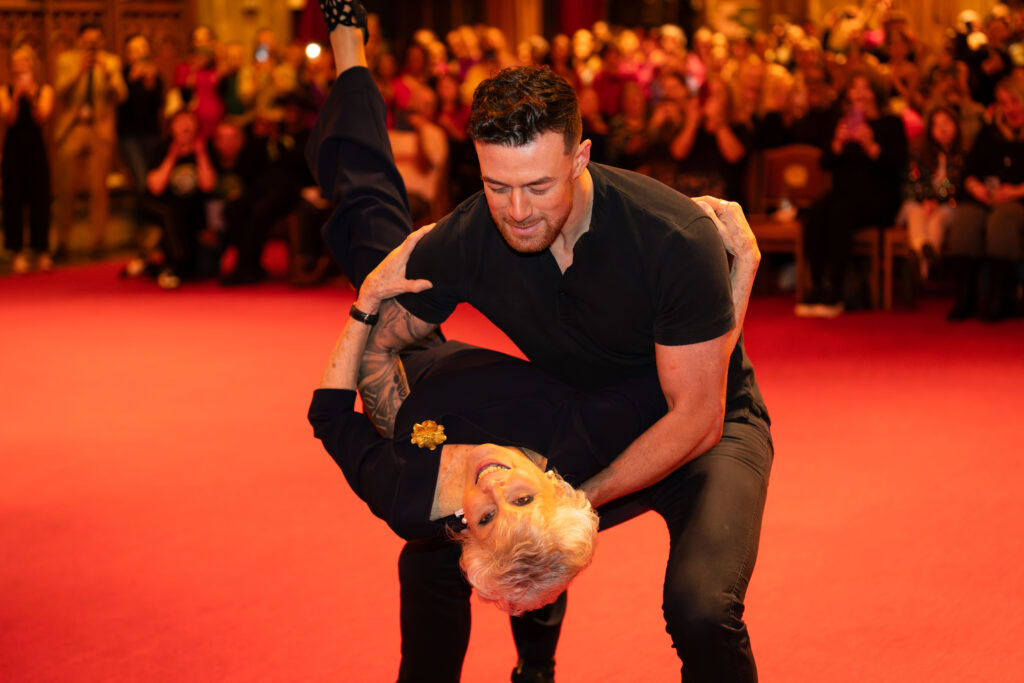 Kai Widdrington dips Angela Rippon while a crowd of onlookers cheer and take photos.