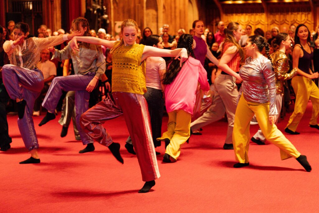 A group of brightly costumed adolescent dancers performer on a red carpet in front of a standing crowd.