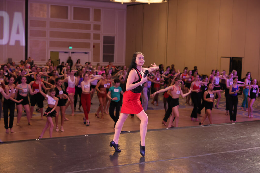 A female ballroom teaching standing on stage leading a ballroom full of young dancers 