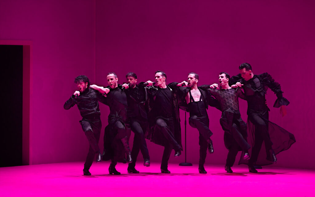 A line of seven male dancers rest their right elbows on each other's shoulders as they hold a mic up to their mouths. Their slightly raise their right legs in front of them as they lean to their right.