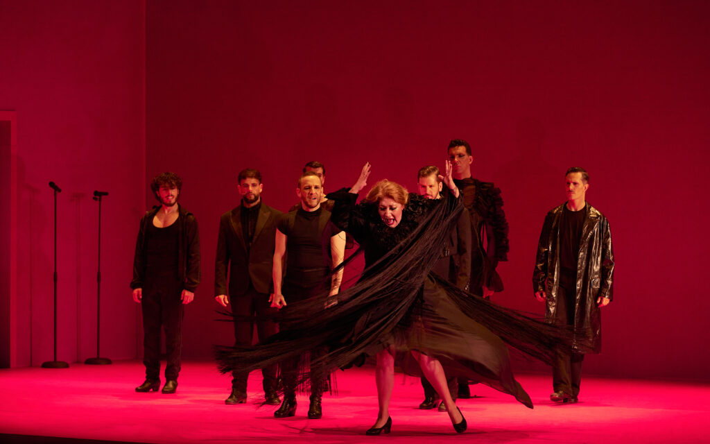 On a red lit stage, seven performers watch intently as downstage, a woman throws her arms up, long black skirts flying around her knees.