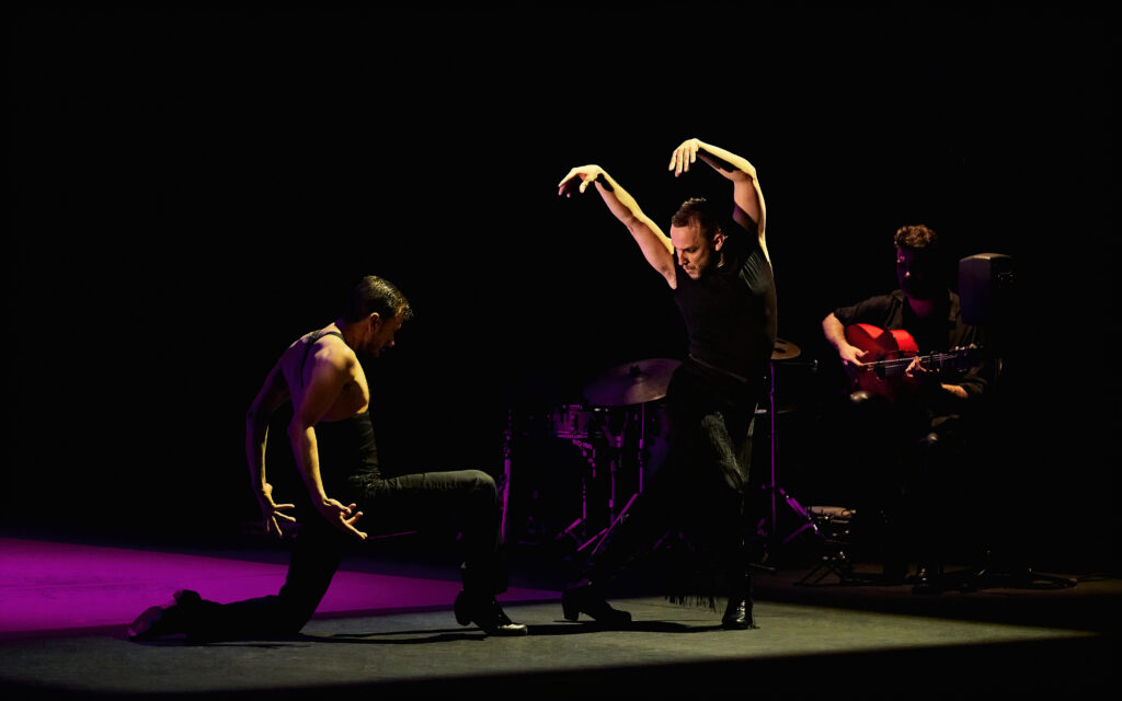 Two dancers on a shadowy stage. One kneels, arms curving behind him and chin lowered, the other twisting to face him with arms curved overhead. Just in the shadows, a seated guitarist strums.