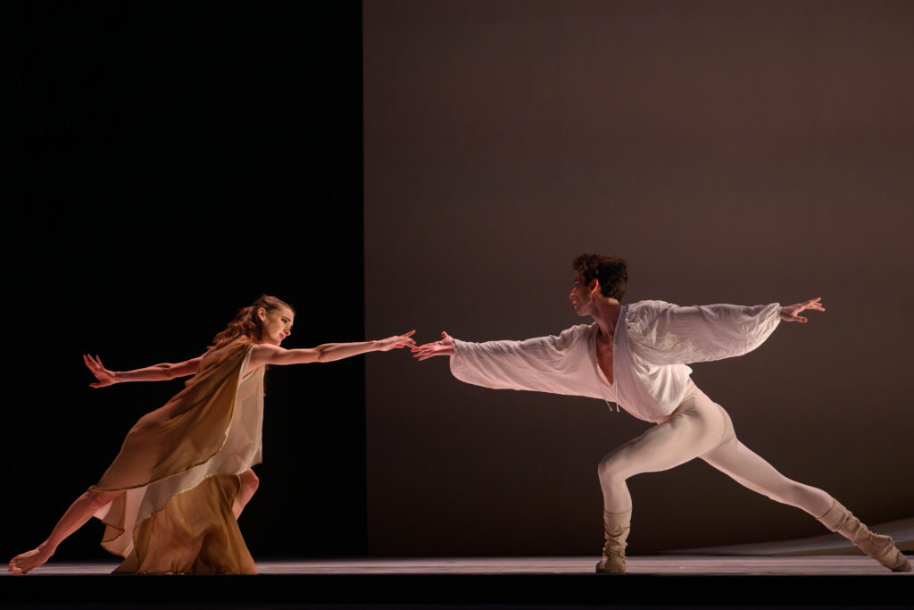 Downstage, two dancers lunge toward each other, arms outstretched so their fingers just brush. One is barefoot and costumed in a golden gown; the other wears a white tunic over white tights and ballet boots.