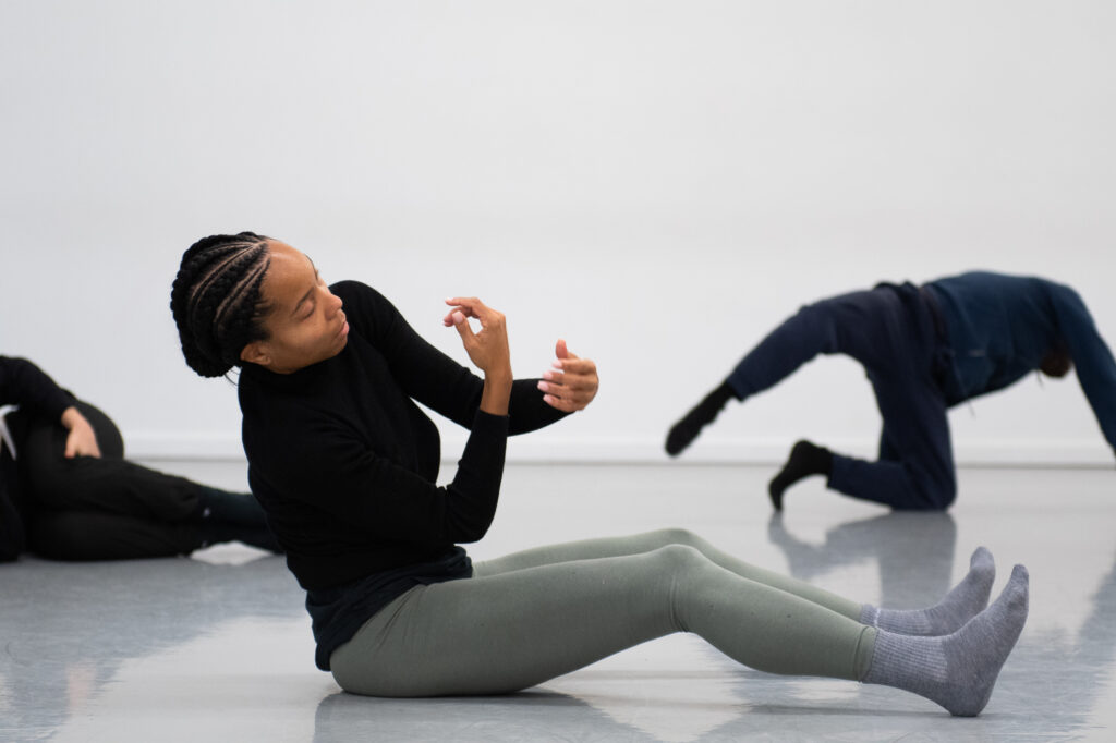 Sidra Bell sitting on the floor in the studio with her legs extended front and her arms curled inward. 