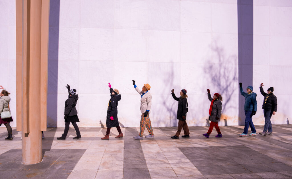 A long single-file line of dancers, wearing a variety of winter coats and accessories, processes in front of the Kennedy Center's white marble exterior. Each dancer is raising their right arm over their head. 