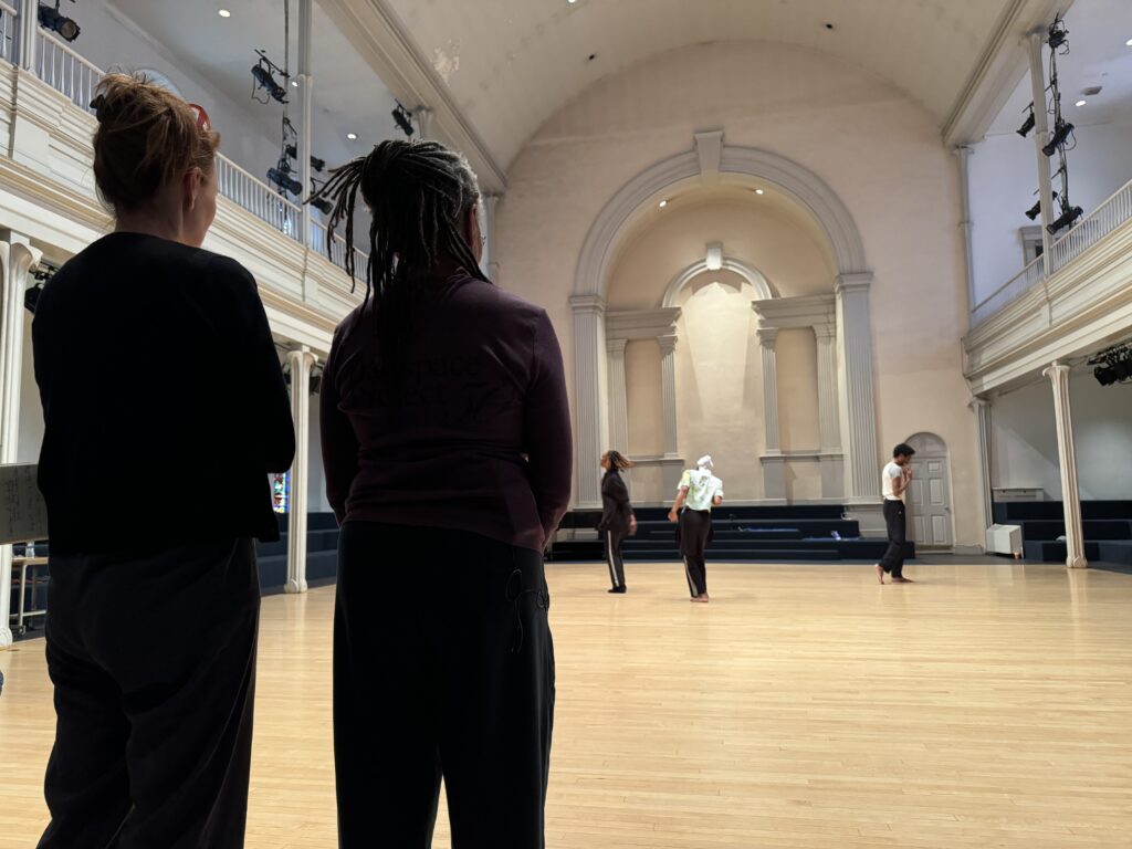 Two figures stand in the foreground with their backs to the camera as they watch a trio of dancers in the Danspace sanctuary.