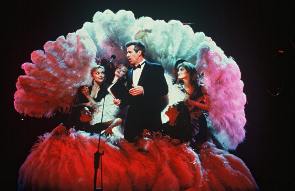 A male performer surrounded by female dancers holding large feather props 