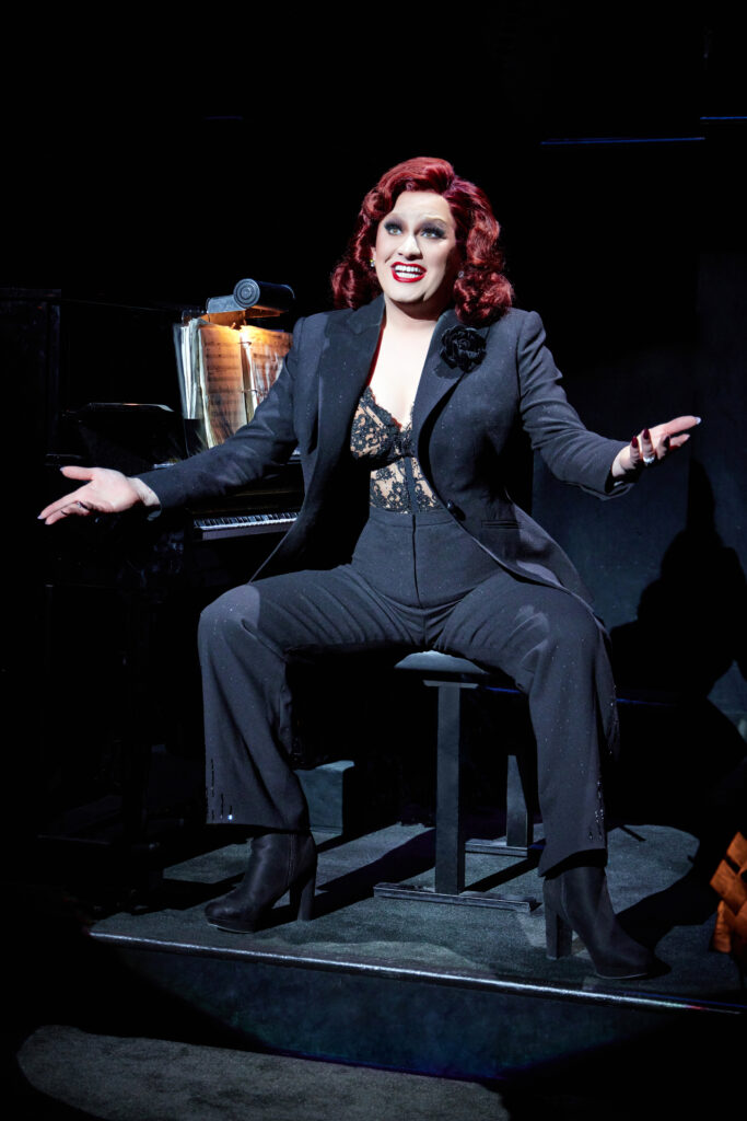 A performer with red hair, lace bodice, and black suit sits on a stool at a piano 