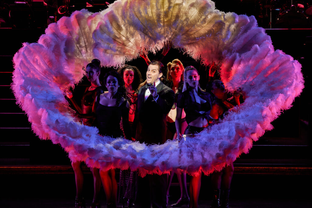 A male performer surrounded by female dancers holding large feather props 