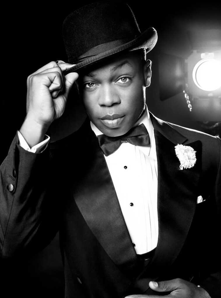 A male performer wearing a suit, hat, and bow tie, tipping his hat at the camera