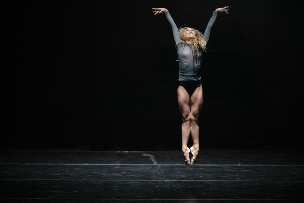 A female dancer on stage jumping straight in the air with her legs tight together and her arms extended up. She wears a blue long-sleeved top.