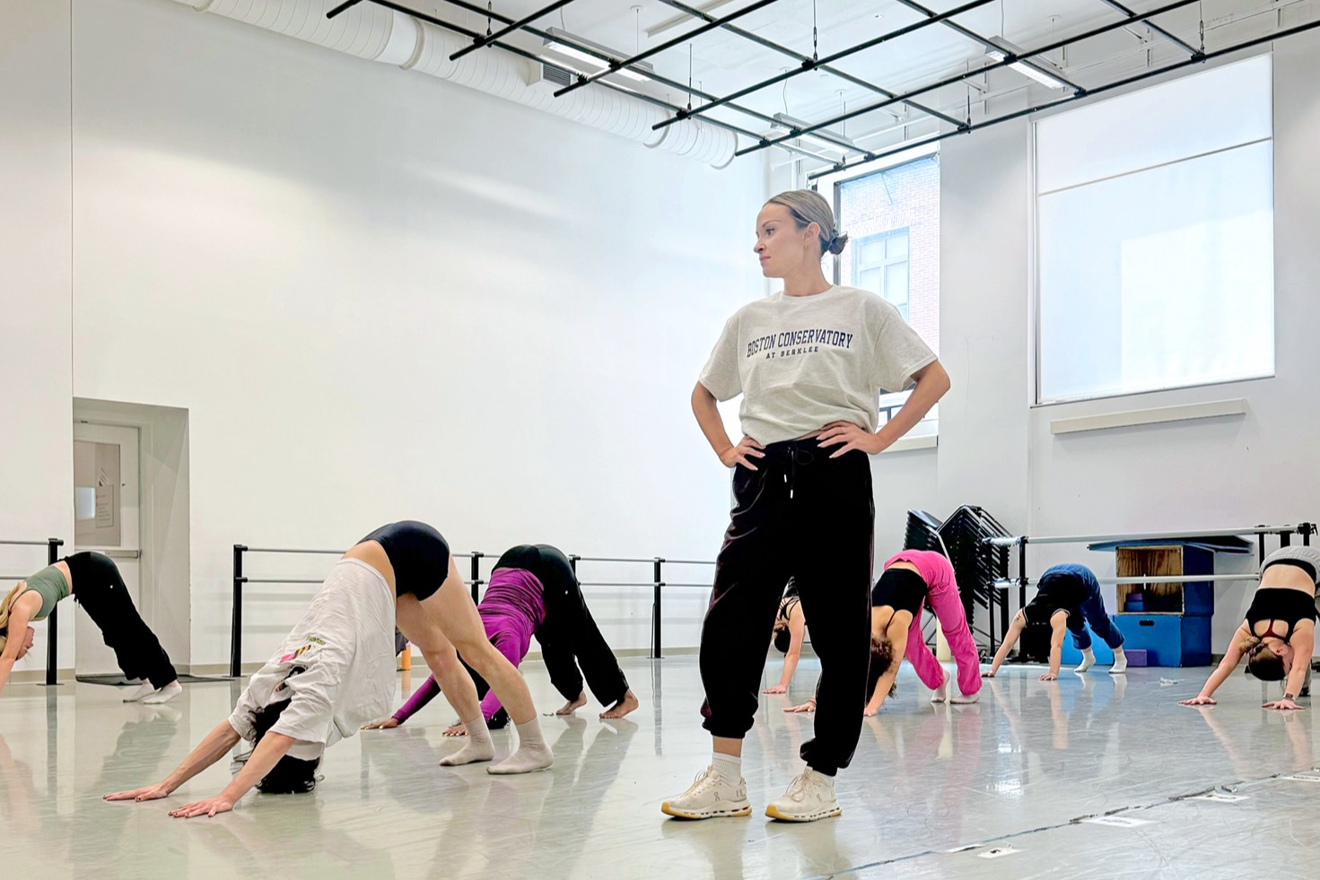 A female instructor standing with her hands on her hips as she watched students stretch in down dog in the studio.