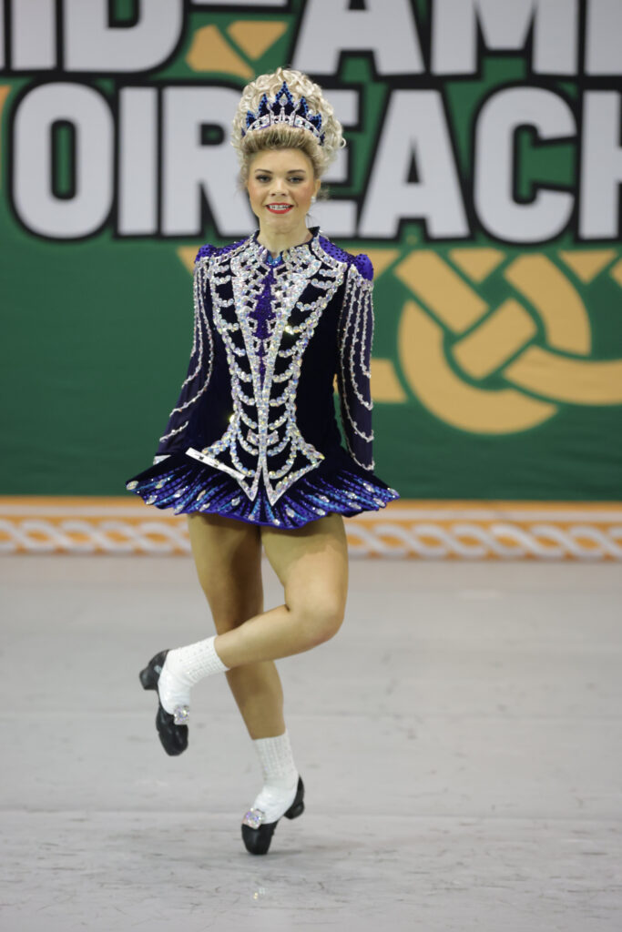 A young Irish dancer mid-step at competition. She wears a detailed deep blue dress and large crown.