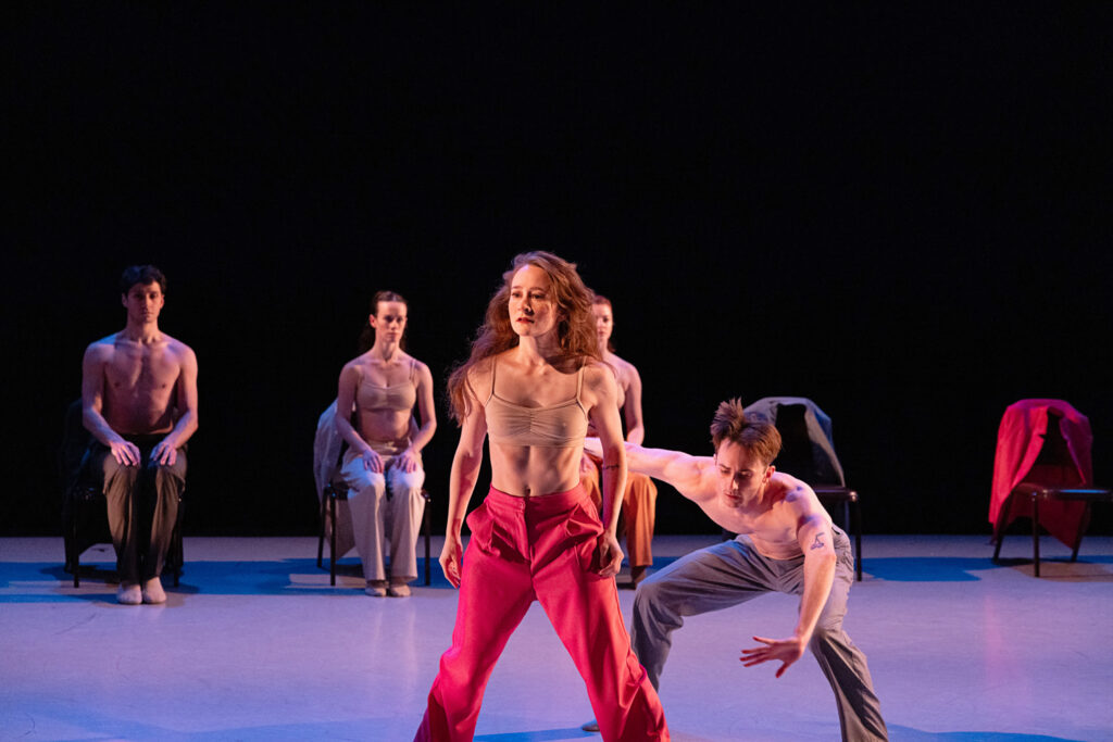 Maggie Carey in a wide stance facing the audience. Yuri Rogers performs a deep plie behind her. Two other dancers sit in chairs upstage. 