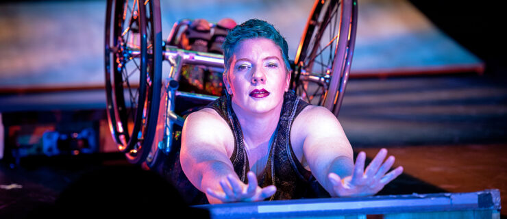 A close-up from Under Momentum. Laurel Lawson lies on their stomach on a low ramp, arms stretching out to the camera in a luxurious arch, wheelchair wheels shining. Lawson is a white person with cropped blue hair and a deep crimson lip. She gazes intently at the viewer. Golden purple light glints from her wheels and creates a glow on her skin.