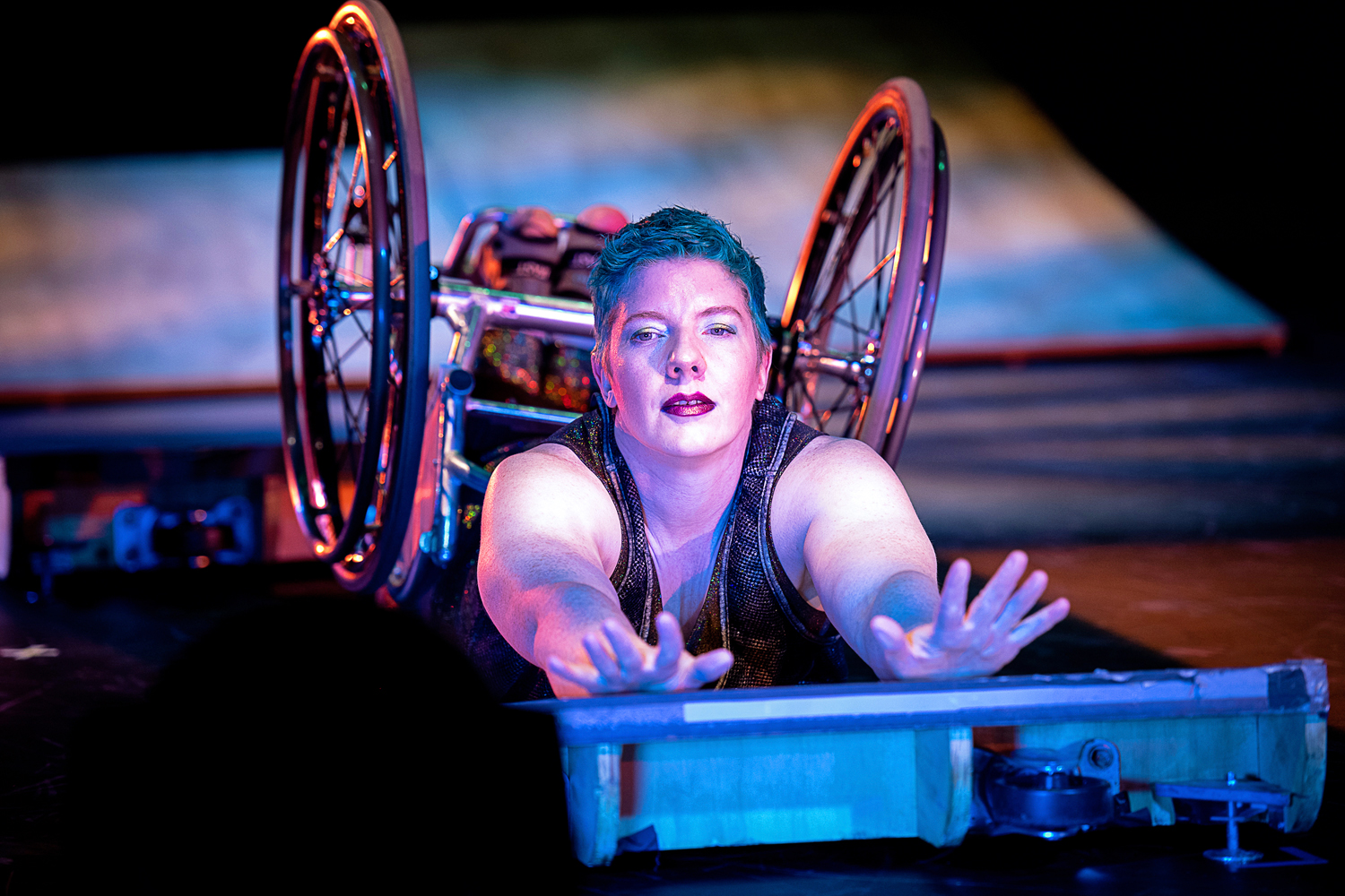 A close-up from Under Momentum. Laurel Lawson lies on their stomach on a low ramp, arms stretching out to the camera in a luxurious arch, wheelchair wheels shining. Lawson is a white person with cropped blue hair and a deep crimson lip. She gazes intently at the viewer. Golden purple light glints from her wheels and creates a glow on her skin.