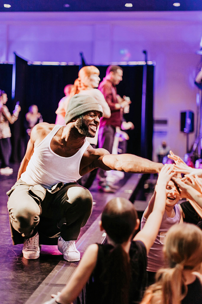 DJ Smart teaching at a convention. He squats on stage and reaches towards the students for a hands-in group cheer. 