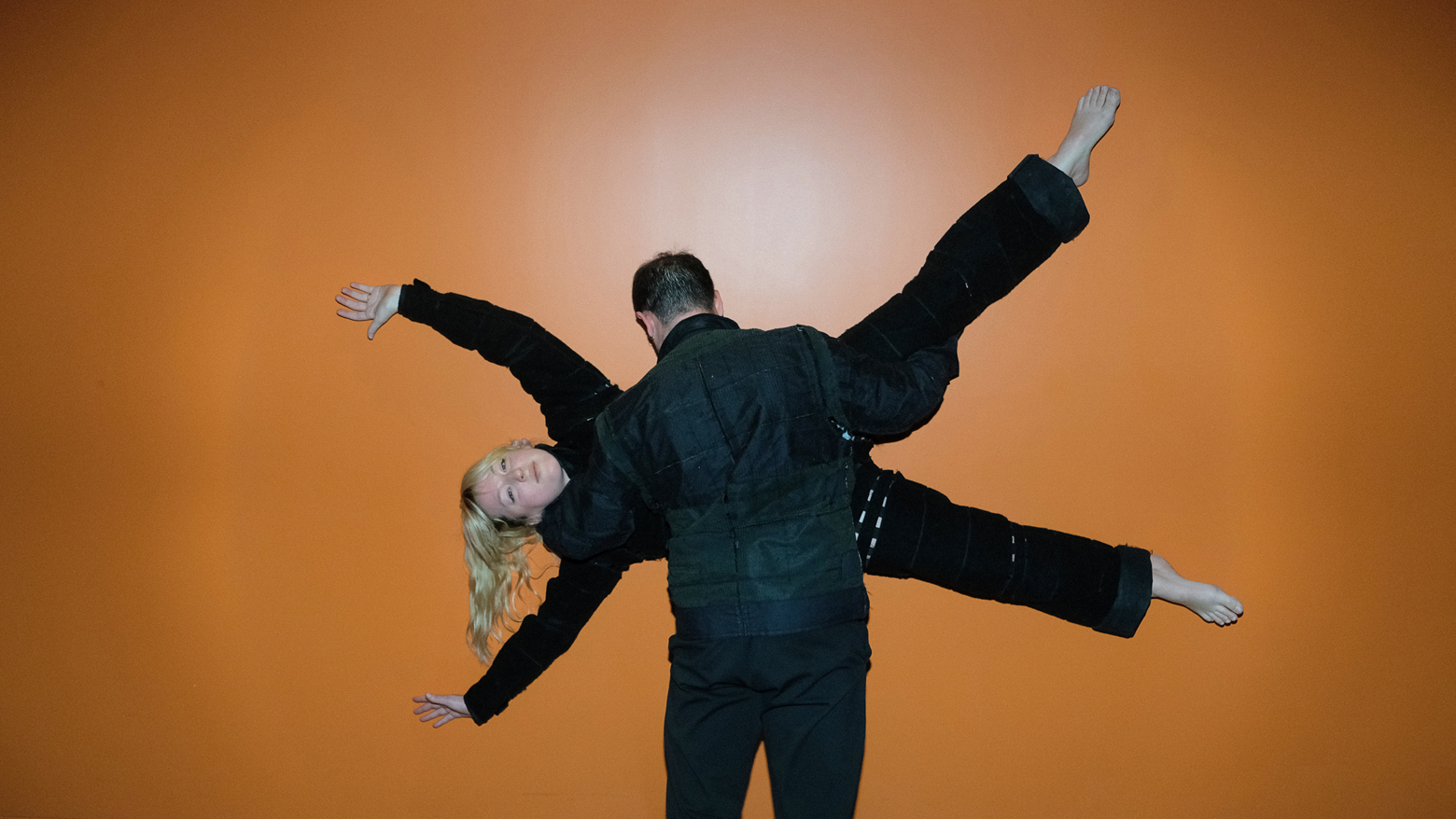 Two dancers partnering together while wearing Velcro-covered suits. One dancer is horizontal with their arms and legs extended out while the other supports them with arms wrapped around.