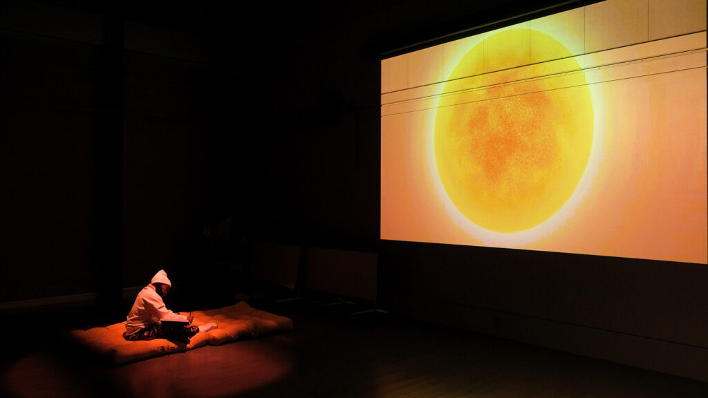Sage Ni’Ja Whitson sitting on the floor onstage in front of a projector showing a bright, large planet.