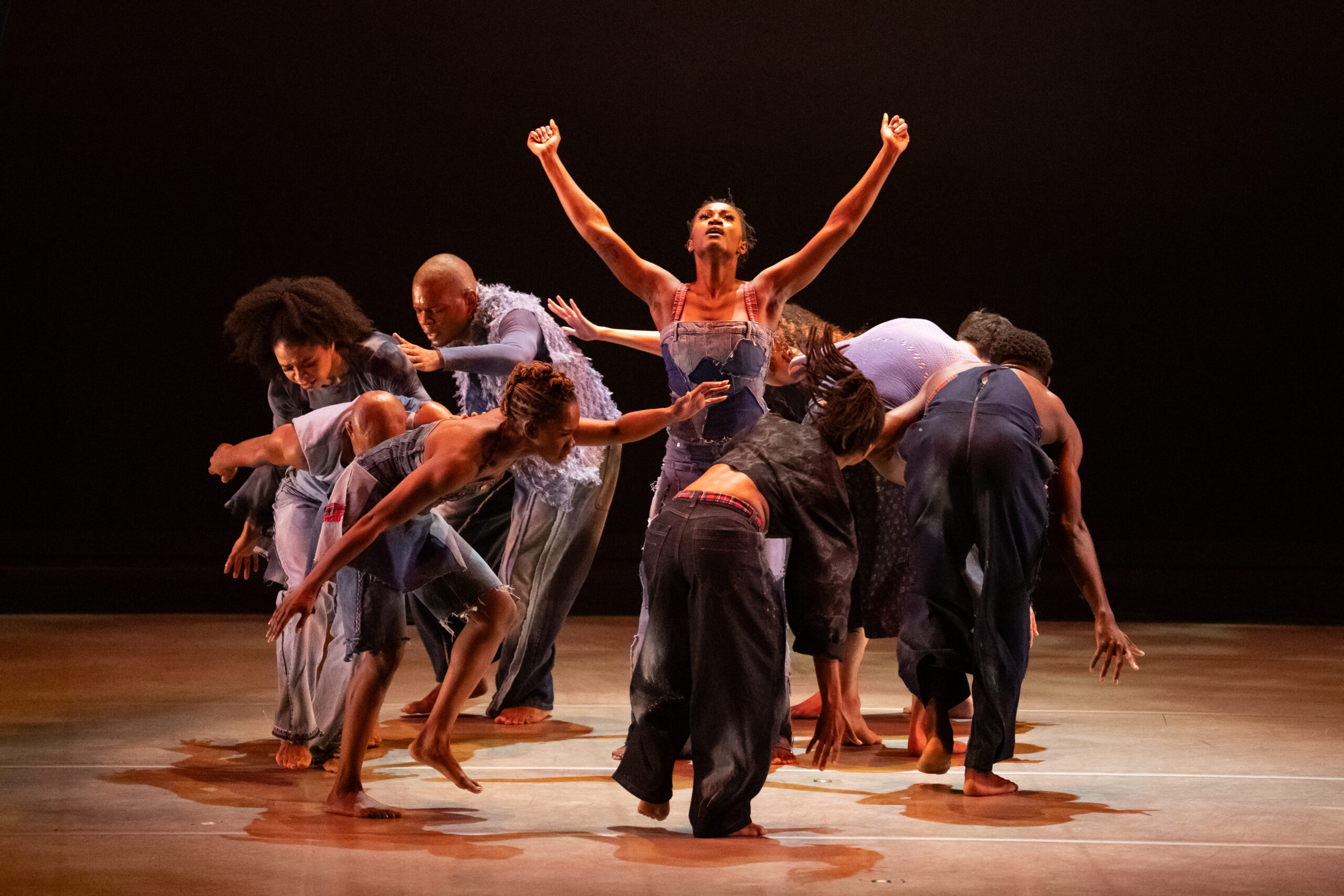 Eight dancers form a circle, bending forward and reaching their left arms for each other's shoulders as they follow each other. A dancer stands at the center of a circle, arms upraised with closed fists. The dancers are barefoot and costumed in lavender, blue denim, and dark grey.