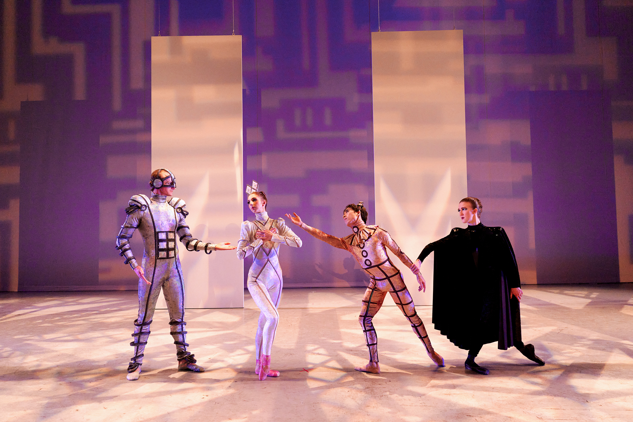 Three ballet dancers onstage dressed as robots, and one as their master. The robots wear silver and gold shiny bodysuits with head pieces. The master wears all black with a large cape.