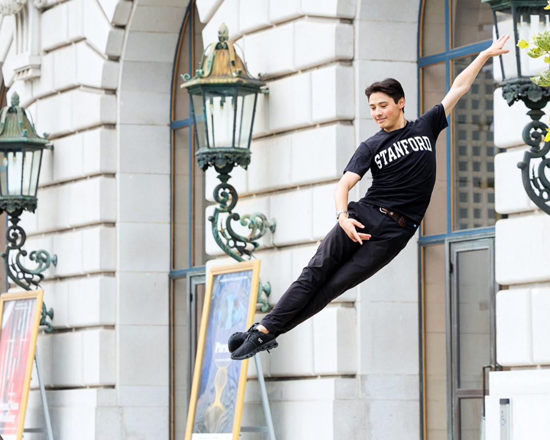 Lleyton Ho jumps in the air with his legs in sous sous. He wears a Stanford t-shirt, black pants, and sneakers.