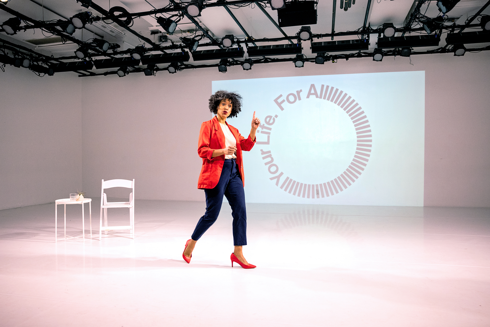 Leslie Cuyjet strides through a white-box theater, gesturing as she speaks. She wears a red blazer and matching heels, a white blouse, and navy pants. On the back wall is projected the words "For All Your Life" styled into a circle with many Ls in the word "all." A white folding chair and a small side table with a glass of water on it are set to one side.