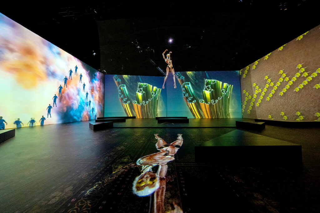 A robot floats in the air in an immersive performance space. On multiple walls and the floor are colorful projections.