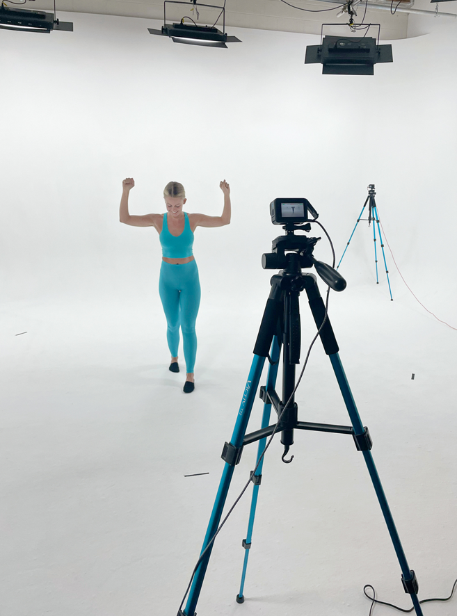 A female dancer wearing a blue athletic set. She moves while standing in front of a white backdrop. Multiple cameras and lights surround her.