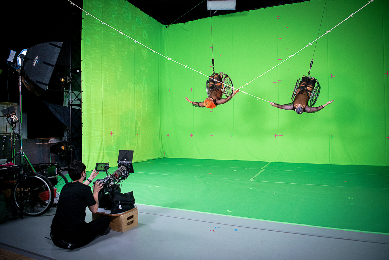 Sheppard and Lawson soar in their wheelchairs against a bright green screen and floor, each holding on to a white barbed wire prop in the shape of a huge X. They are both upside down, faces tipped and arms curved toward the ceiling. Lauren Mendoza sits on the floor in the foreground, with a large camera propped up on a wooden box, filming the dancers from below. Alice is a multiracial Black woman with short bright orange curly hair and coffee-colored skin; Laurel is a white person with pale skin and cropped peacock blue hair. They wear copper bodysuits overlaid with black mesh.