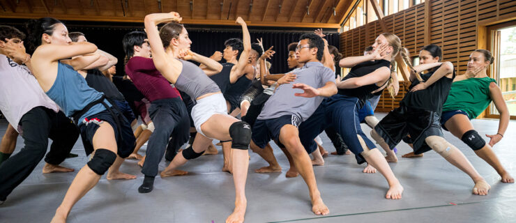 A large group of male and female presenting dancers in multi colored athletic clothing gather in clump facing each other. They each lean their upper body backwards while in a wide stance with their legs bent and far apart, and bring their hands up to create a shape of their own. The image portrays a powerful and confrontative energy.