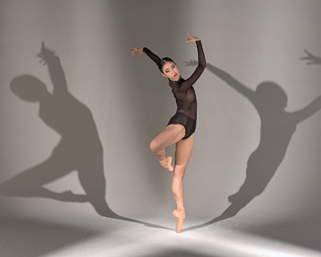Agnes Su dances against a photo backdrop wearing pointe shoes and a black, long sleeve leotard. She performs a retiré en pointe and casts her shadow along the wall.