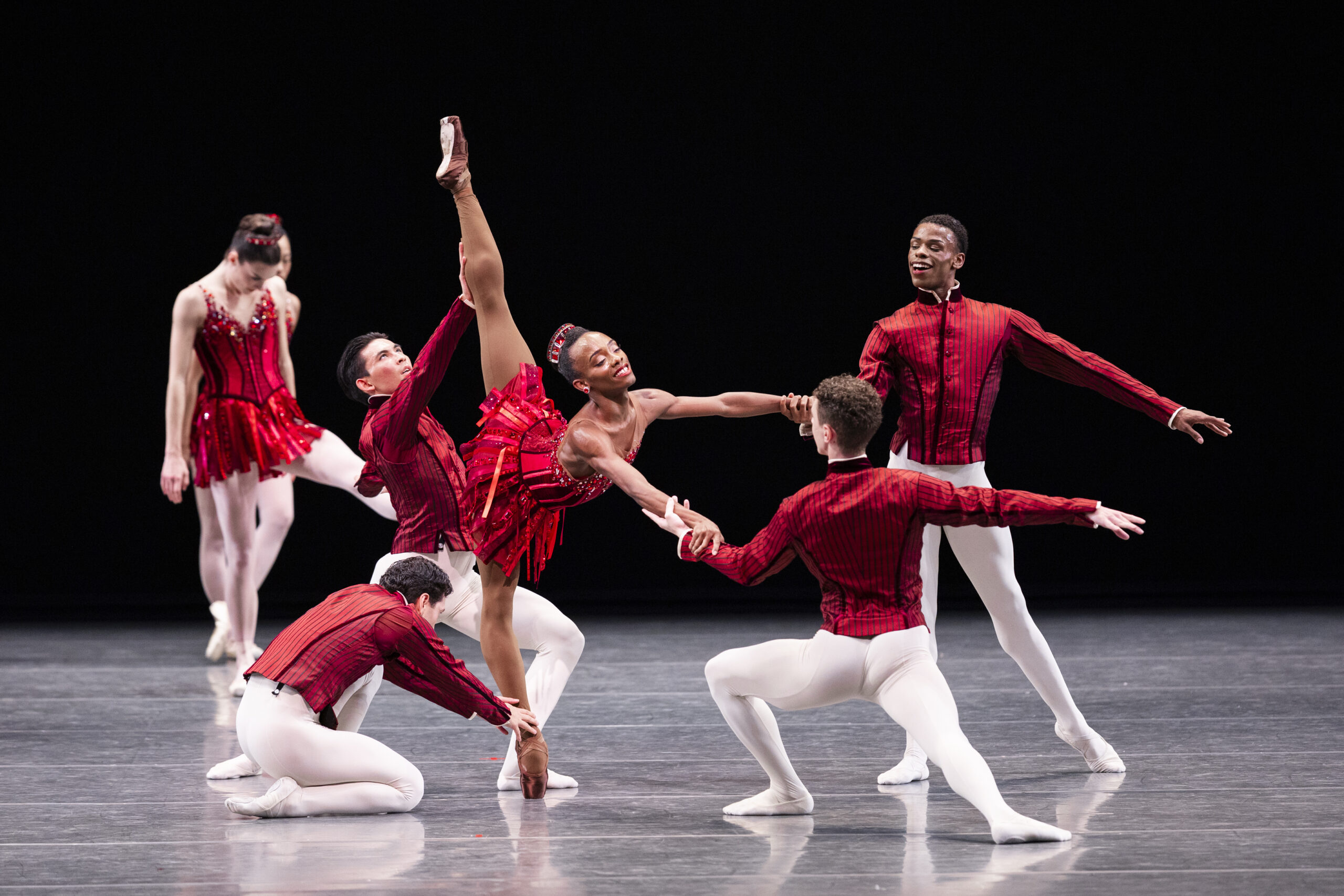 Ashton Edwards in a supported penché on pointe, supported by a dancer at each of their limbs. They are costumed in a red dress and tights and pointe shoes that are pancaked to match their skin tone. The dancers around them wear red long-sleeved tunics and white tights.