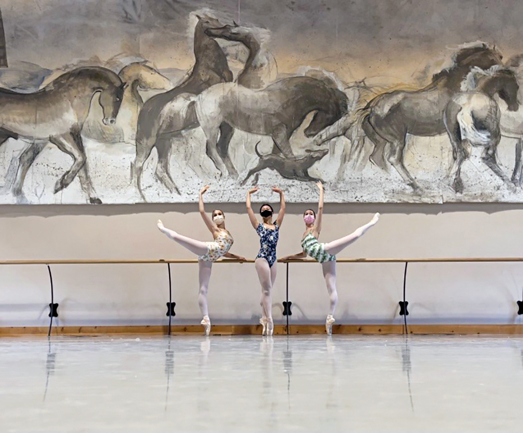 Three dancers at the barre in a studio with a large mural on the walls. They perform an attitude derrière en pointe with the dancer in the middle in sous-sus.