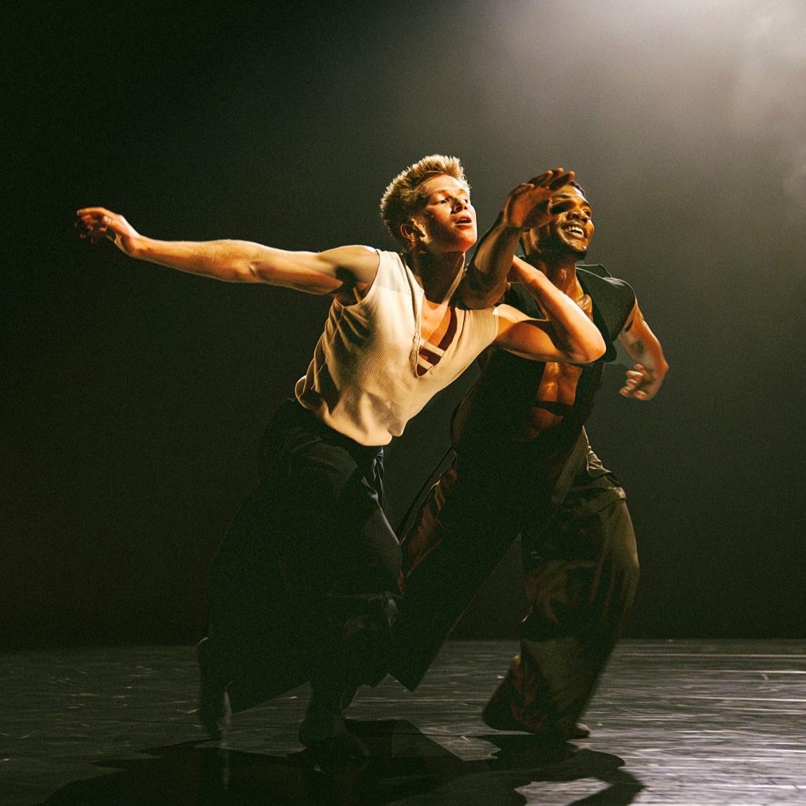 Joziah German (left) and Sidney Chuckas dancing on a dimly lit stage. They swing their arms and lean forward.