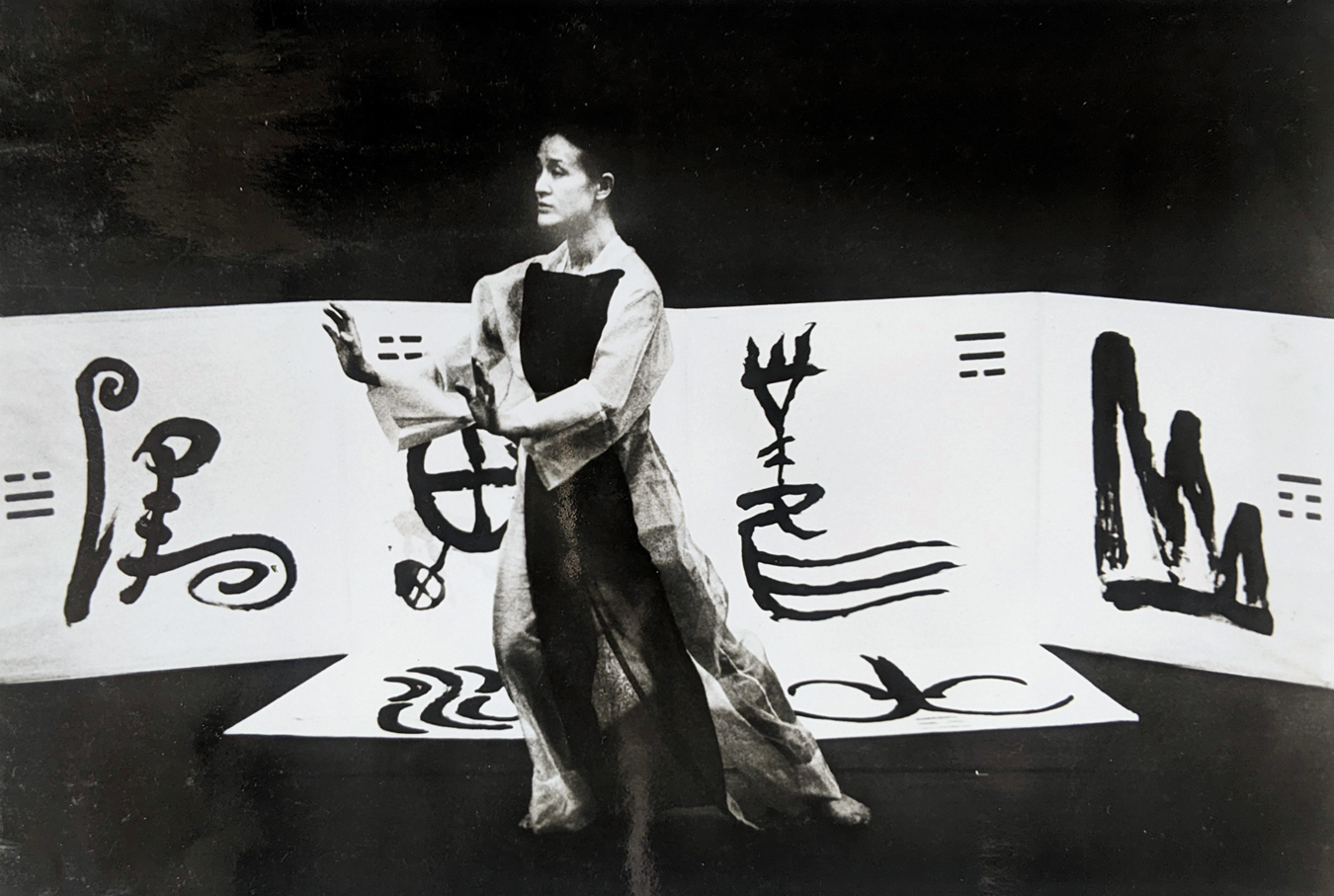 A black and white photo of Chiang Ching performing. She wears a long flowy dress and dances in front a white backdrop with symbols.