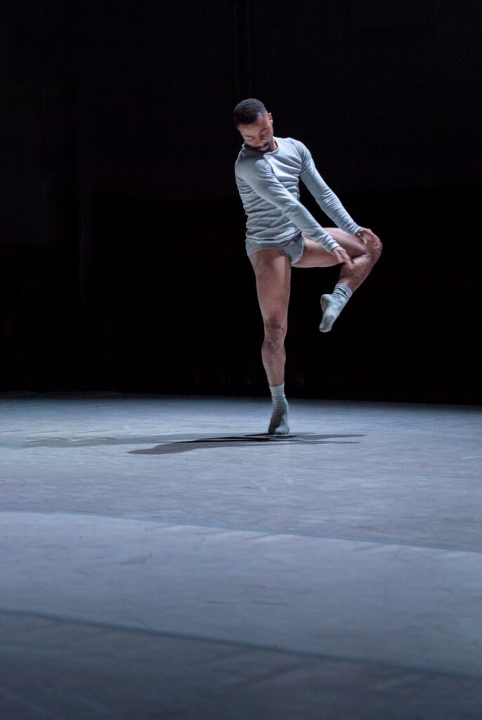Roderick George balances on relevé, arms stretched down toward his upraised knee. He looks down over his shoulder toward his standing leg.