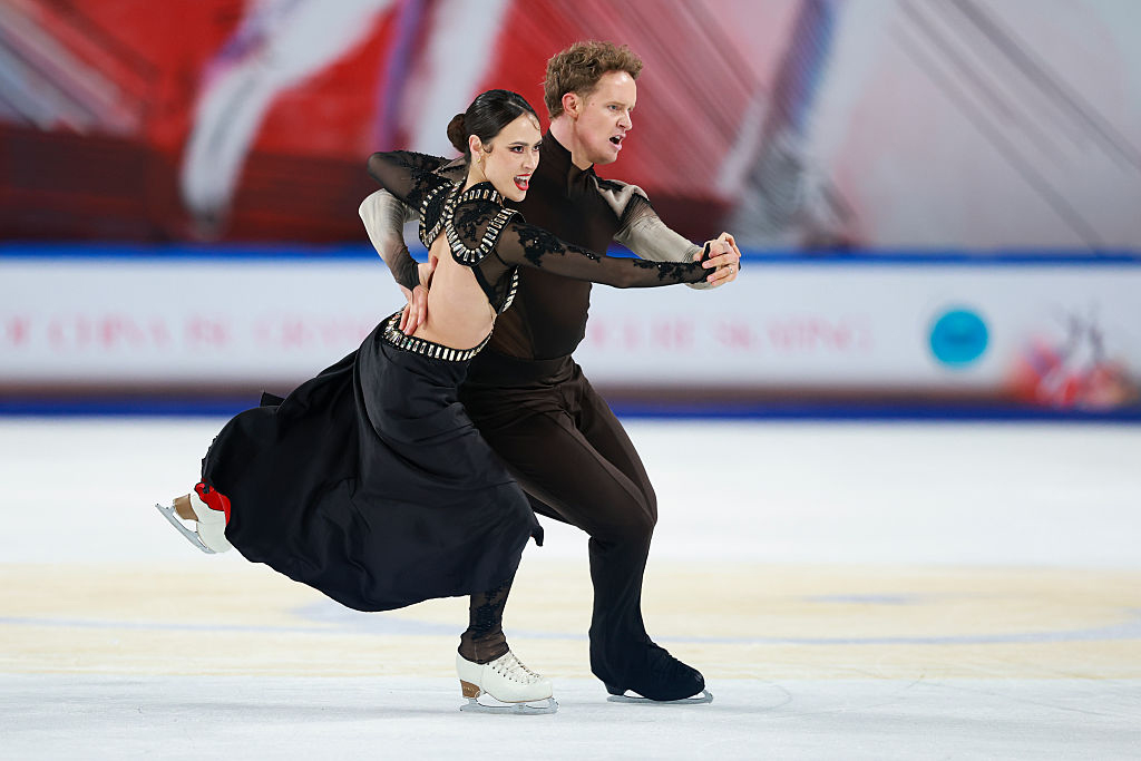 Chock and Bates, in black costumes accented in red and gold, are captured at an intense moment in their ice dancing routine, their arms clasped as they charge forward together.