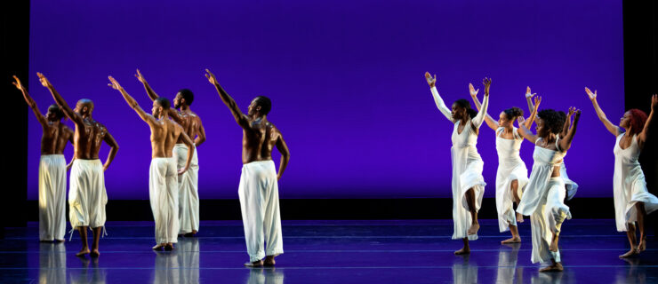 Five male dancers and five female dancers perform in two clusters on either side of a purple-lit stage. The male dancers, bare-chested and costumed in white pants, face an upstage diagonal, left arms raised on a high diagonal as they look offstage. The female dancers, dressed in flowing white blouses and pants, lift their arms overhead, palms upturned, as they raise a flexed foot by their calves. Their chins are upraised as they look toward the men.
