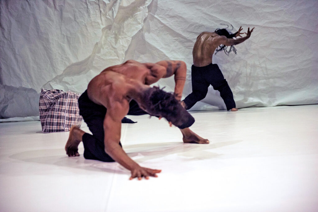Two dancers throw themselves forward, one slamming a hand to the ground and the other raising his arms just shy of the crumpled white backdrop. Both are barefoot and bare-chested, and wear black pants.