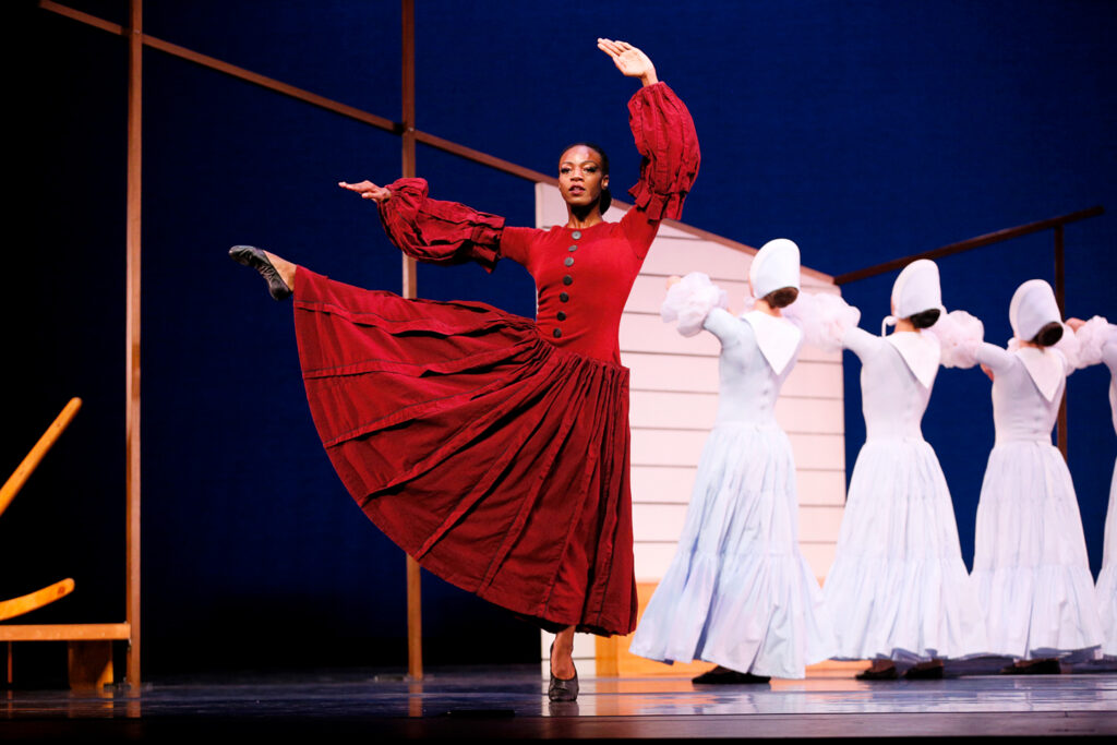 Leslie Andrea Williams balances on relevé with a leg extended side, opposite arm curved overhead with a flat palm facing out to the audience. She is costumed in a long red dress with billowing sleeves and black slippers. Behind her, a line of dancers in similar dresses in pure white face upstage in a line. The sparse set forms the outline of a house.