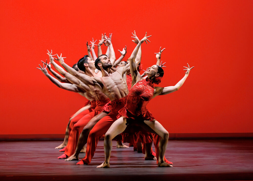 A line of dancers in second position plié stretches upstage. Their arms curve overhead, fingers splayed, as they lean back and twist toward the center; alternating dancers lean left or right. The backdrop is lit red to match the dancers' costumes.