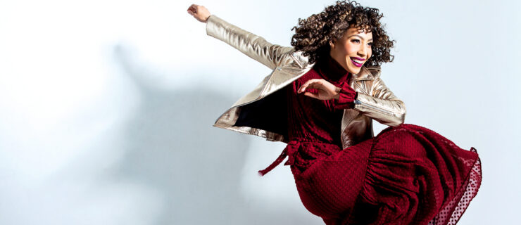 Afra Hines swings her arms while lifting her right knee the opposite direction. She wears a long red dress, a shiny jacket, and sneakers.