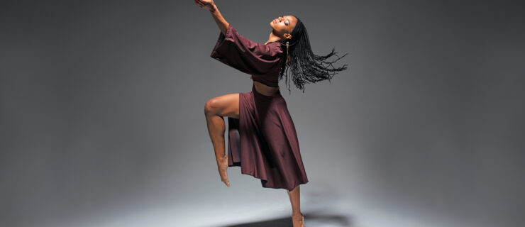 Leslie Andrea Williams falls forward off of relevé, right knee raised in front of her with a pointed foot. Her opposite arm rises in front of her, palm upturned as though to receive something from above. Her face is tipped up to the light, eyes closing. Her braids fly in the air behind her. She is barefoot and wears a maroon skirt and cropped shirt with draping sleeves.