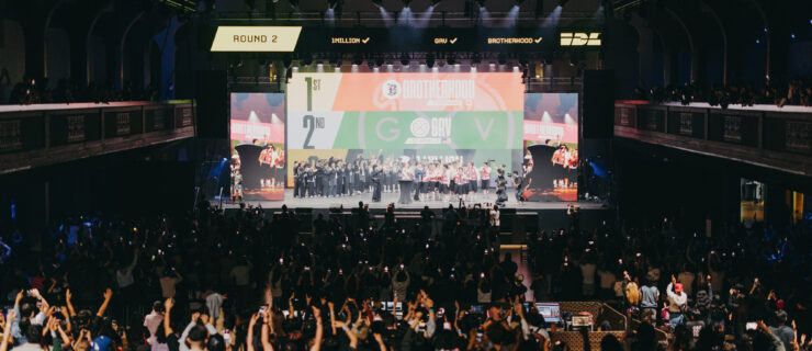 In a large, dark arena, a crowd cheers a group of dancers on a raised stage.