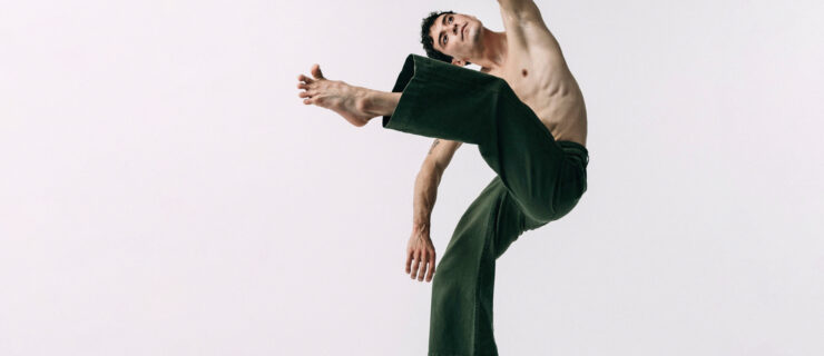 Jacob Beasley stands on one foot en relevé while lifting the other leg in attitude front and leaning laterally the same direction.