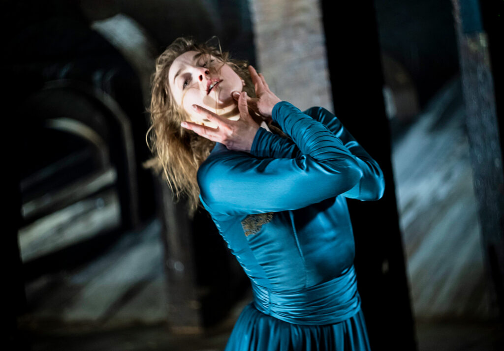 A dancer in a formal blue dress crosses her arms at the elbow to bring her palms to her face. Her hair is loose down her back as her chin tips up.